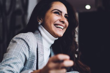 Kahkahalarla gülen, uzun siyah saçlı, günlük kıyafetlerle neşeli, kafede dizüstü bilgisayarla oturup notlar alan bir kadın.