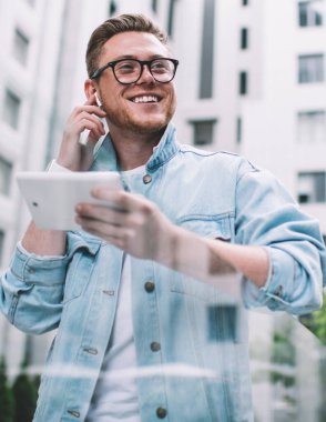 Kot ceketli, kulaklıklı, modern tablet kullanan ve sokakta mutlu bir şekilde gülümseyen mutlu bir adamdan.