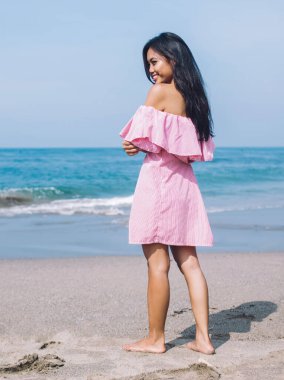 Arkaya bakan pembe elbiseli Endonezyalı genç kadın güneşli bir günde okyanusun arka ucunda gülümsüyor.