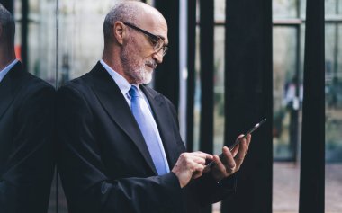 Sokak binasının duvarına yaslanırken, resmi kıyafetli, gözlüklü ve gözlüklü, sakallı, yakışıklı ve kel bir adamın yan görünüşü.