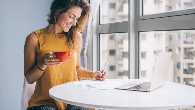 Sarı kolsuz bluzuyla beyaz yuvarlak masada oturan, evinde kağıt üzerinde kırmızı kupa yazan mutlu genç bir kadın.