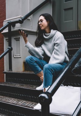 Kot pantolon ve rahat süveter giyen güzel yetişkin bir kadın New York 'un caddesinde gezerken alışveriş çantalarını merdivenlere koyuyor.