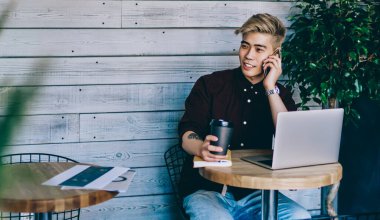 Çinli hippi öğrenci elinde kahve ile modern kafenin iç kısımlarında dizüstü bilgisayarda oturuyor. Japon genç adam cep telefonuyla konuşurken bilgisayar başında serbest çalışıyor.