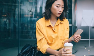 Modern elbiseli düşünceli bir Asyalı kadın sırt çantasıyla oturuyor ve Hong Kong 'da cep telefonuyla sörf yaparken kahve içiyor.