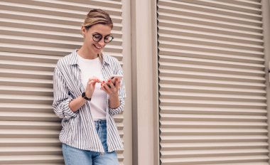 Genç ve güzel bir kadın günlük kıyafetler giyiyor ve moda gözlükler takıyor. Gülümsüyor, akıllı telefon kullanıyor arkadaşlarıyla sohbet ediyor ve gri pencereye yaslanıyor.