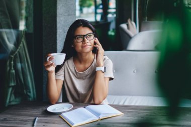 Gözlüklü genç ve çekici bir kadın kafede defterle otururken telefonda konuşuyor güzel bir kadın kahve molası sırasında elinde bir fincan sıcak içecekle telefon görüşmesi yapıyor.
