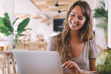 Akıllı telefon ve belgelerle masada rahat bir koltukta oturan, laptopunu kullanan ve bulanık arka planda, restoranda hindistan cevizi suyunun tadını çıkaran hoş bir esmer kadın.