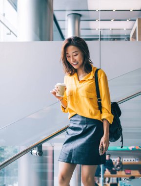 Hong Kong 'da kahve ve cep telefonuyla merdivenlerden aşağı inen sarı tişörtlü, siyah deri etekli ve sırt çantalı güzel, pozitif, güzel Asyalı kadın.