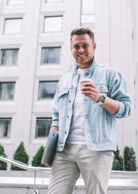 Sokağın ortasında kameraya gülümseyen, tek kullanımlık kahve ve dizüstü bilgisayarlı yakışıklı genç adamdan.