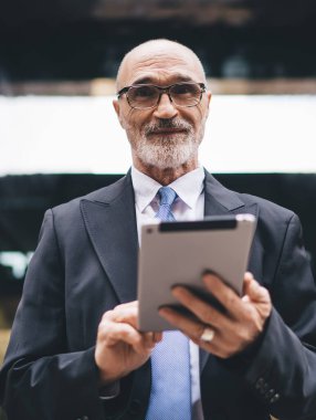 Yakışıklı, sakallı, kel, resmi kıyafetli ve gözlüklü bir adam iş merkezinin önünde kameraya bakıyor.