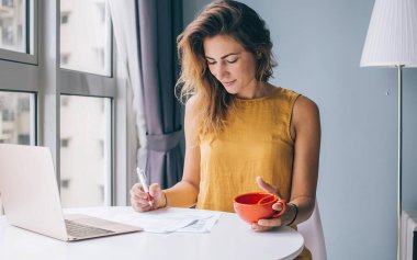 Konsantre olmuş, sarı kolsuz bluzlu, beyaz yuvarlak masada oturan, laptoplu ve kırmızı kupalı, evde kağıda yazılmış genç ve çekici bir kadın.