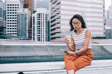 Çağdaş, gözlüklü, modaya uygun kıyafetleri olan, kahve içip tablet seyreden Hong Kong görüşlü genç bir kadın.