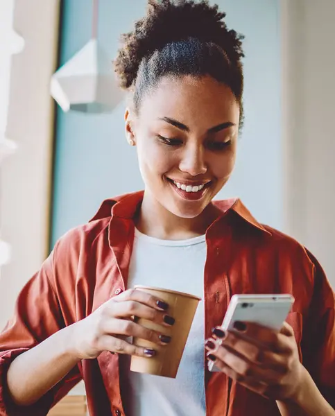 Happy dark skinned hipster girl sending sms message via smartphone device while enjoying caffeine beverage, african woman holding paper cup with coffee and reading article on content funny website