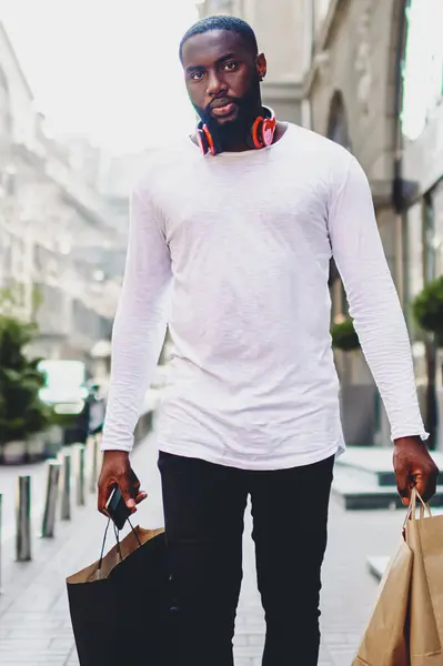 Portrait of handsome afro american guy carrying paper bags walking on city street after shopping, dark skinned male in long sleeve shirt with copy space for brand name or label strolling outdoors