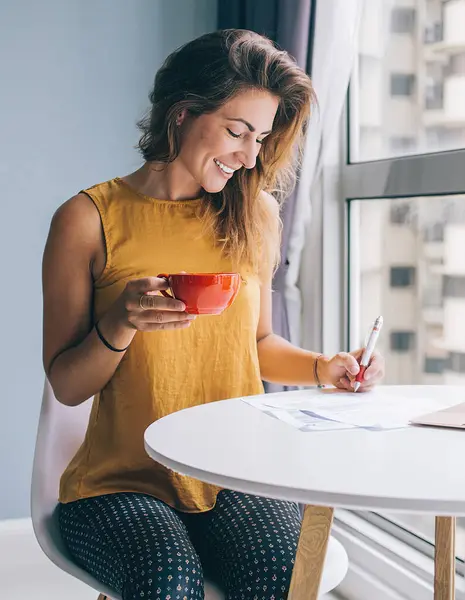 Delighted young attractive woman in yellow sleeveless blouse sitting at white round table with laptop and red mug writing on paper at home