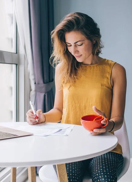 Concentrated young attractive woman in yellow sleeveless blouse sitting at white round table with laptop and red mug writing on paper at home