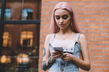 Dövmeli, uzun pembe saçlı, kot elbiseli modern odaklı kadın. Şehir caddesinde cep telefonundan mesajlaşıyor.