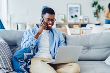 Neşeli Afro-Amerikan hipster adam cep telefonuyla konuşuyor. Evden internet üzerinden uzaktan çalışıyor. Gülümseyen erkek serbest yazar, dizüstü bilgisayardan bankacılık servisini arıyor.