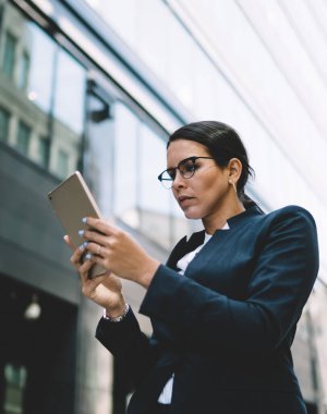 Gözlüklü, siyah saçlı, resmi takım elbiseli, tablet giyen ve şehirde gündüz yürürken ekrana dikkatle bakan bir kadın.