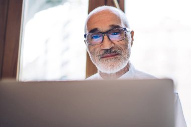Beyaz gömlekli, mavi kravatlı yaşlı, kel, sakallı yönetici. Pencerenin yanındaki bulanık arka planda bilgisayar kullanıyor.