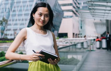 Elinde not defteri olan, modern ofis binasının balkonunda dikilirken kameraya bakan şık kıyafetli pozitif etnik yönetici.