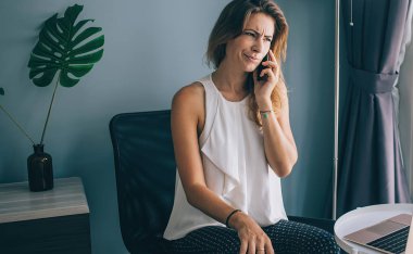 Kafası karışmış genç esmer, gündelik giysiler içinde masanın yanındaki koltukta dizüstü bilgisayarla oturuyor ve akıllı telefonla konuşuyor.