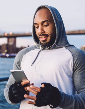 Genç Afro-Amerikalı adam ayakta durup kulaklıkla müzik dinlerken akıllı telefondan mesaj atıyor ve nehir ve köprünün arka planına hafifçe gülümsüyor.