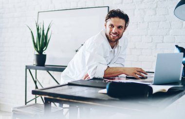 Neşeli Kafkas erkek serbest çalışan, modern tasarlanmış iş yerinde uzaktan kumandalı iş yapan, başarılı gülümseyen iş adamı, ofisteki bilgisayarın yanında online bir iş için dikiliyor.