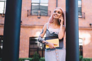 Not defterleri ile mutlu genç bir bayan. Gülümsüyor ve gözlerini kaçırıyor. Bir yandan da direğe yaslanıyor ve telefon çağrılarına cevap veriyor.