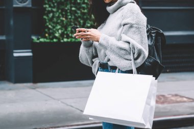 Modern yetişkin kadın, günlük kıyafetleri ve gözlükleriyle elinde alışveriş çantası ile New York 'un caddesinde yürürken akıllı telefonuna göz atıyor.