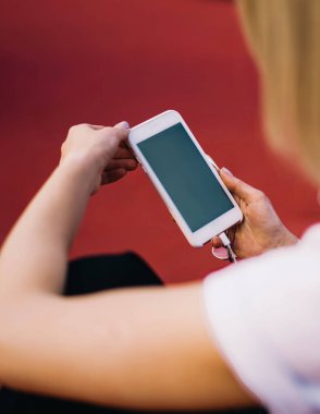 Arka plan tanınamaz halde. Moda kıyafetleri içinde, lastik spor sahasında otururken mesaj göndermek için cep telefonu kullanan bir kadın.