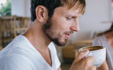 Güzel milenyum erkeğine odaklanın. Sıcak, keskin kahvenin tadını çıkarırken arka planda bulanık bir kadın kağıda yazı yazıyor ve gülümsüyor.