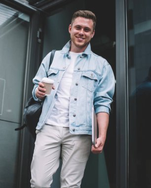 Kot ceketli, elinde bir fincan kahve ve dizüstü bilgisayarla caddede kameraya gülümseyen mutlu genç adamdan.