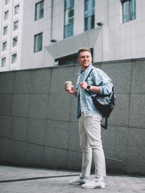 Rahat giyimli, sırt çantalı, elinde tek kullanımlık kahveyle sokakta durup gülümseyen havalı bir genç.