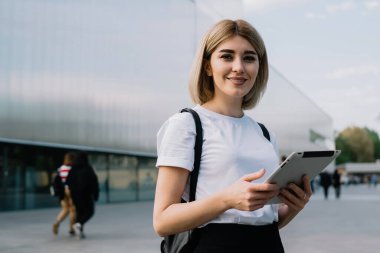Günlük kıyafetlerle gezen neşeli kadın, şehir caddesinde ofis binasının yanında durup kameraya gülümserken zevkle tableti karıştırıyor.