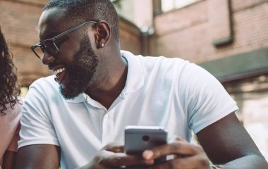 Gözlüklü, gülümseyen hippi, akıllı telefon ekranına bakarak sevgili kız arkadaşının yazdığı SMS mesajlarını kontrol ediyor. Aşık çift akıllı telefon cihazlarında 4G kablosuz internet kullanıyor.