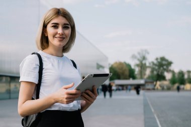 Günlük kıyafetlerle gezen neşeli kadın, şehir caddesinde ofis binasının yanında durup kameraya gülümserken zevkle tableti karıştırıyor.