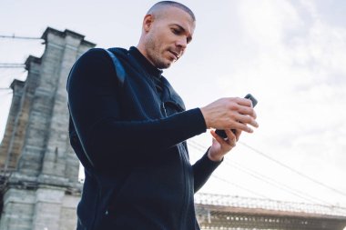 Aşağıdan keyifli, traşsız, siyah spor giysili genç adam şehir köprüsünde güpegündüz metal çitlerde akıllı telefonları dinliyor.