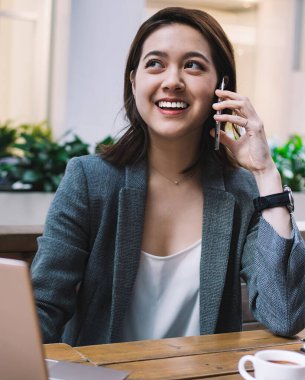 Resmi olarak gülümseyen Asyalı serbest çalışan bir kadın bilgisayarı incelerken bir kupa kahve ve not defteriyle uzaktan çalışıyor.