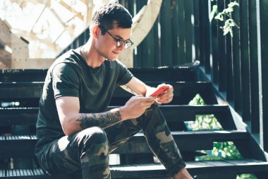 Haki kıyafetli, akıllı, genç, sıska bir adam yazın şehirde merdivenlerde oturuyor.