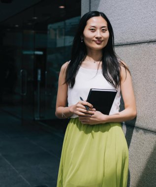 Keyifli stil sahibi genç Asyalı kadın gündüz vakti şehir ofis binasının yanında dururken günlük kıyafetlerle organizatör planlaması yapıyor.