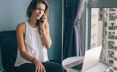 Kafası karışmış genç esmer, gündelik giysiler içinde masanın yanındaki koltukta dizüstü bilgisayarla oturuyor ve akıllı telefonla konuşuyor.