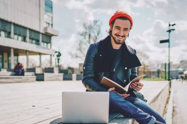 Üniversite sınavına hazırlanmak için vaktini harcayan neşeli beyaz erkek öğrencinin portresi kampüste dizüstü bilgisayar ve eğitim kitabıyla oturuyor, not defteri olan pozitif hipster bir adam.