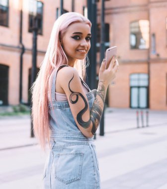 Pembe saçlı, dövmeli, akıllı telefon taşıyan ve sokakta dikilirken kameraya gülümseyen neşeli genç bir kadın.
