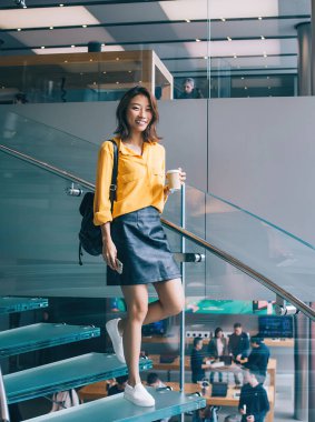 Tam vücut genç, pozitif, güzel, neşeli Asyalı, sarı tişörtlü, siyah deri etekli, sırt çantalı, Hong Kong 'da kahve ve cep telefonuyla merdivenlerden inen bir kadın.