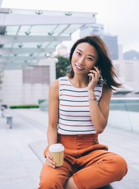 Plastik bardak ve bacak bacak üstüne atmış Asyalı güzel bir genç kadın telefonda konuşurken bankta oturuyor ve Hong Kong 'un arka planındaki kameraya bakıyordu.