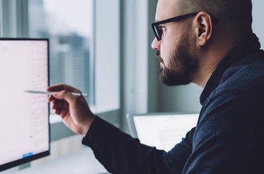 Ofisteki masadaki istatistiksel bilgilerle çalışan, gözlüklü sakallı adamın ekran görüntüsü.