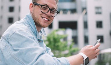 Gözlüklü gülümseyen genç bir adamın yan görüntüsü. Sokakta cam çitlere yaslanmış cep telefonu tutuyor ve kameraya bakıyor.