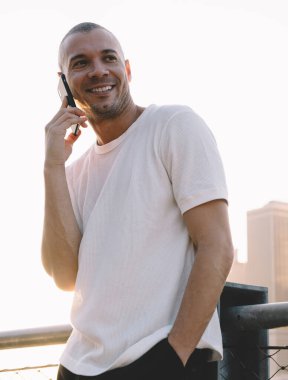 Aşağıdaki beyaz tişörtlü, kısa saçlı adamdan tırabzana yaslanmış ve telefonda gülümseyen.