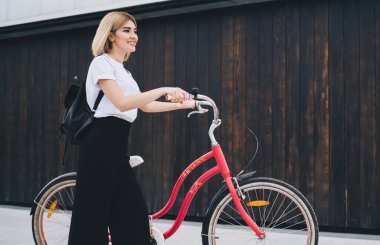Sıradan kıyafetli, siyah sırt çantalı ve bisikletle cadde binasına bakan neşeli, genç, modern sarışın kadın.